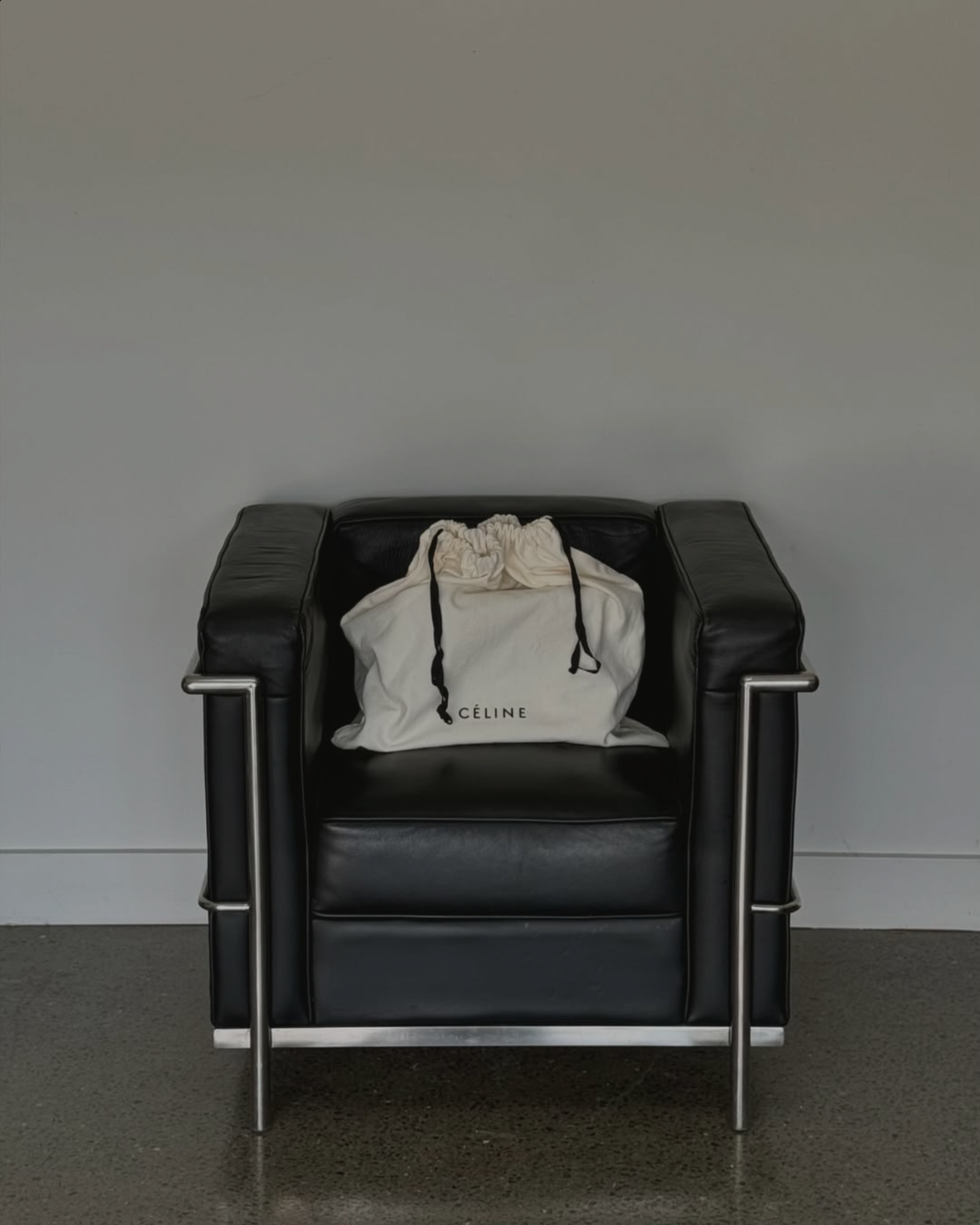 A black leather armchair with a chrome metal frame sits against a plain gray wall on a concrete floor. A cream-colored Celine dust bag rests on the chair.