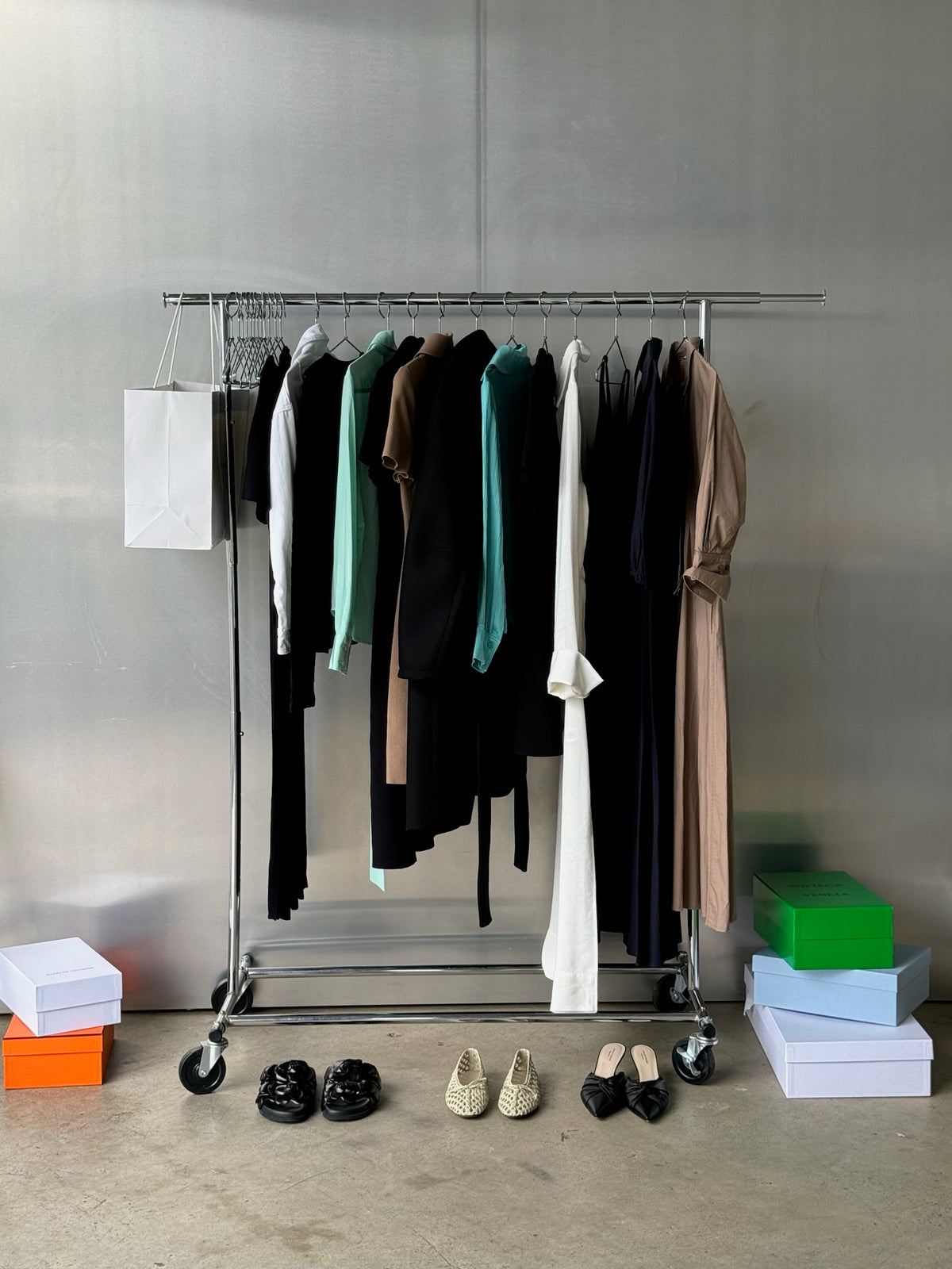 Neutral clothing hung-up on a silver clothing rack against a chrome wall. Shoes and shoe boxes are placed on the floor. 