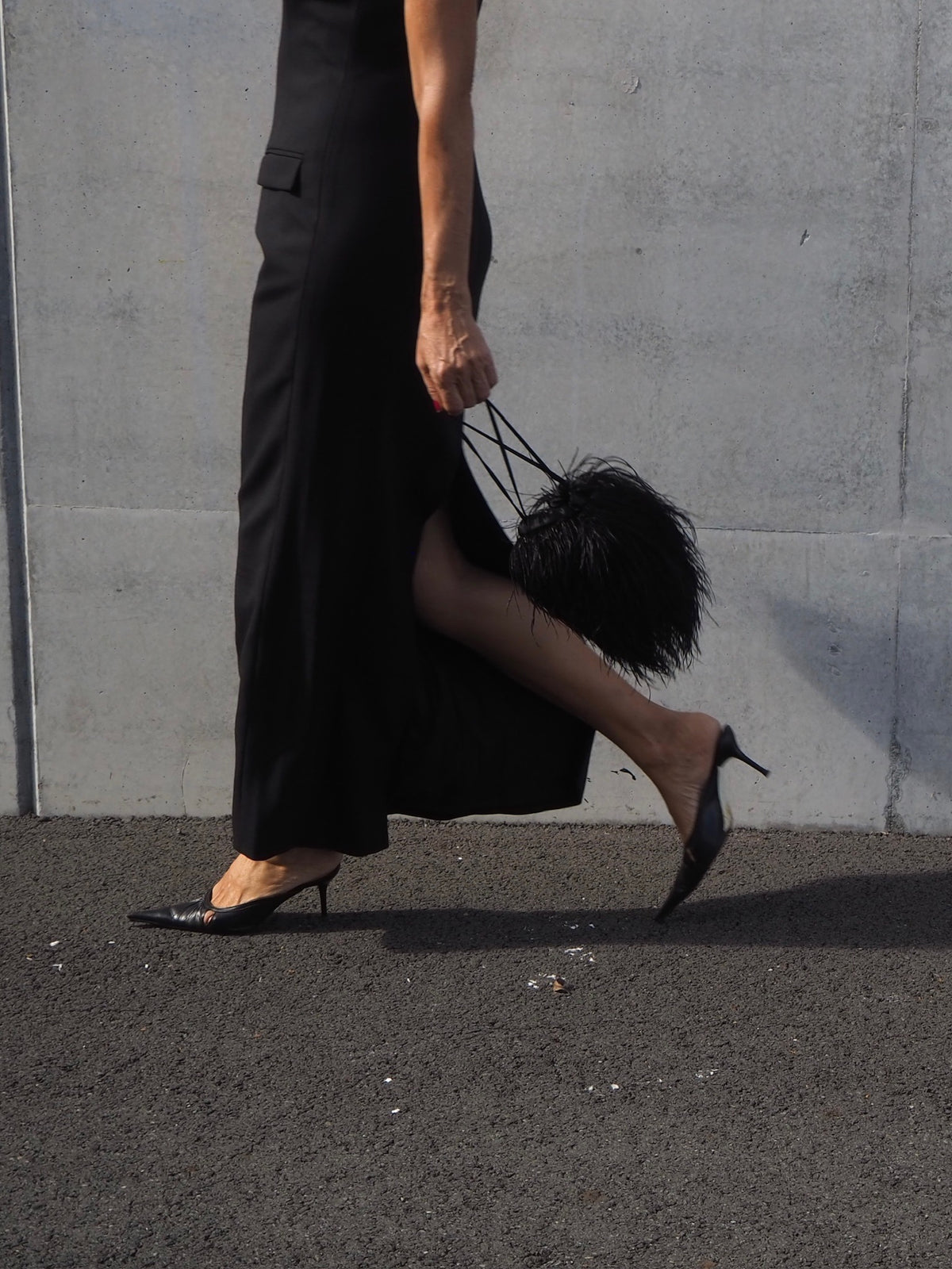 Person wearing a black outfit with high heels and a black handbag against a gray wall.