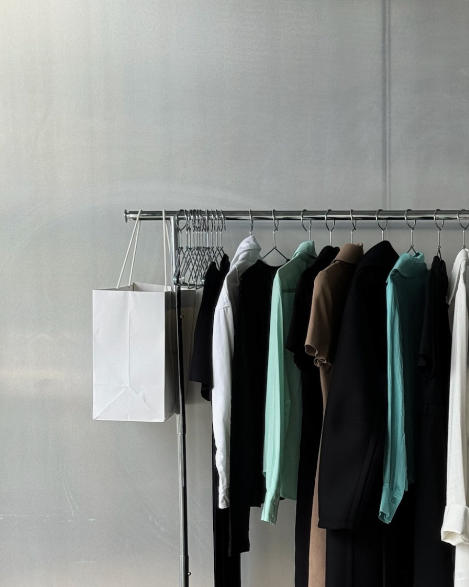 A clothing rack against a light gray wall, holding multiple garments in black, white, mint green, brown, and teal tones, with a white shopping bag hanging on the left.
